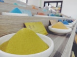 A series of small ceramic dishes filled with colorful powders are lined up on wooden shelves. Each dish contains a different colored powder, including yellow, blue, and orange. There are small labels in front of some dishes with writing on them. The setting appears to be indoors with soft, diffused lighting and a slightly out-of-focus background.