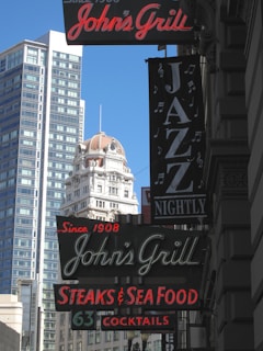 Several vertical neon signs are displayed on the exterior of a building in an urban setting. The signs advertise John's Grill, featuring steaks, seafood, cocktails, and nightly jazz performances. In the background, there are high-rise buildings, including an older ornate structure with a dome.