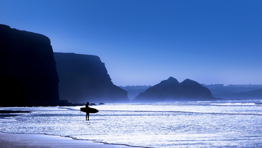 silhouette of person holding surfboard standing on seashore at blue hour