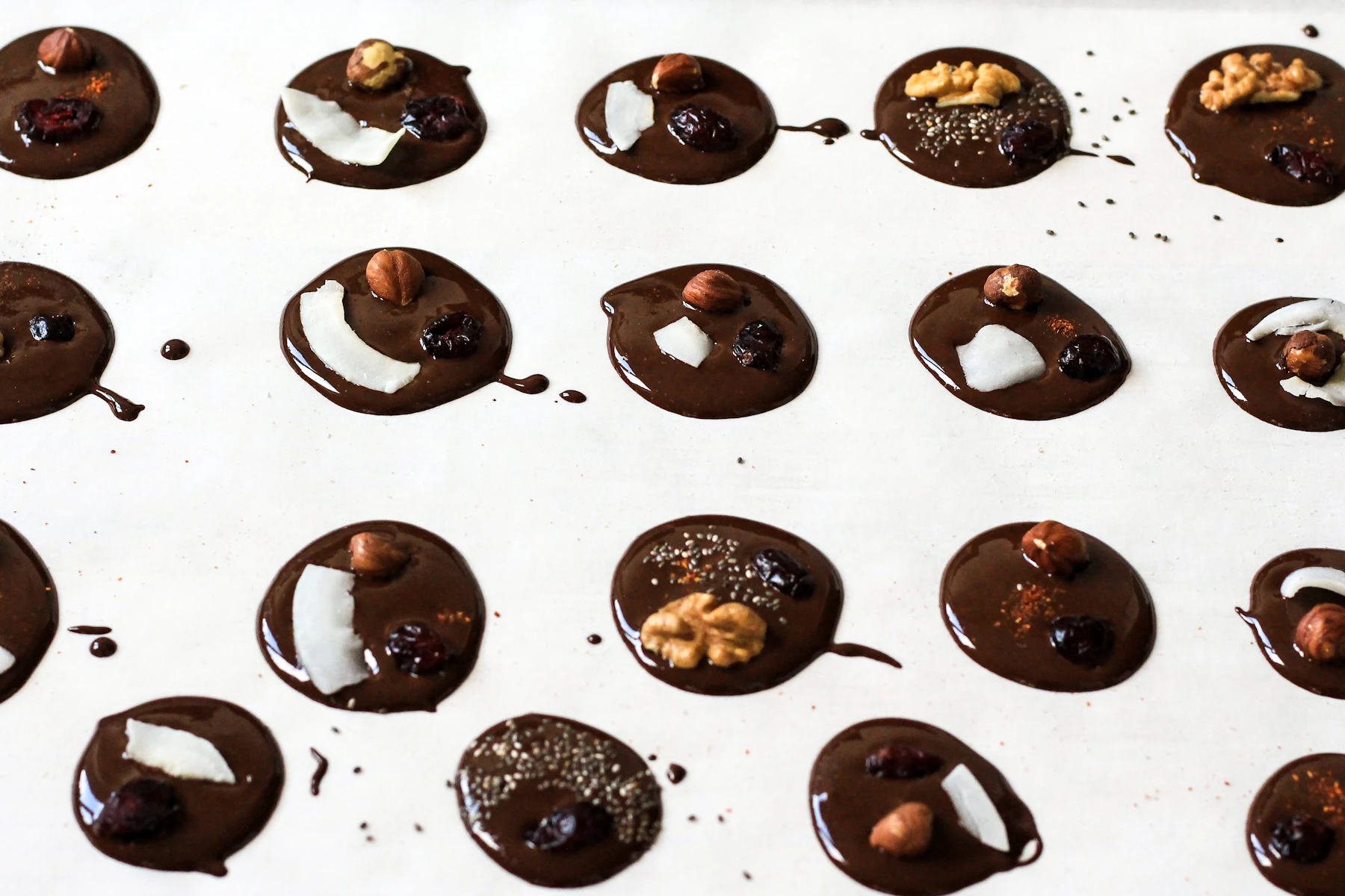 Rows of spooned dark chocolate discs on parchment, topped with nuts, coconut and dried fruit