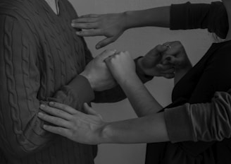 A monochrome image featuring multiple hands in various poses. The focus is on the interaction between the hands and arms, suggesting movement or communication. The texture of a cable-knit sweater is visible on one of the individuals.
