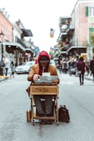 A person sits in the middle of a bustling street with a typewriter placed on a wooden stand labeled 'poet for hire'. The individual wears a red hoodie and a dark hat, with their head lowered, appearing focused on writing. The street is lined with buildings, and flags are visible in the background, contributing to a lively urban atmosphere.