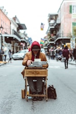 A person sits in the middle of a bustling street with a typewriter placed on a wooden stand labeled 'poet for hire'. The individual wears a red hoodie and a dark hat, with their head lowered, appearing focused on writing. The street is lined with buildings, and flags are visible in the background, contributing to a lively urban atmosphere.