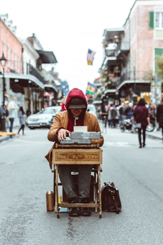A person sits in the middle of a bustling street with a typewriter placed on a wooden stand labeled 'poet for hire'. The individual wears a red hoodie and a dark hat, with their head lowered, appearing focused on writing. The street is lined with buildings, and flags are visible in the background, contributing to a lively urban atmosphere.