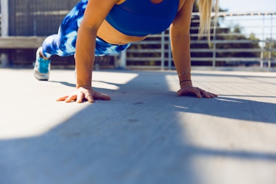 A person is performing a plank exercise on a concrete surface outdoors. The focus is on the person's arms and core, wearing a blue sports bra and blue patterned leggings. Sunlight casts shadows on the ground, and there is a metal railing and trees in the background.