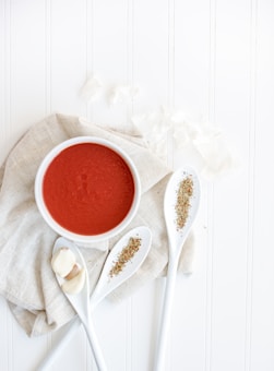 A flat lay composition featuring a white bowl filled with rich red tomato soup resting on a light-colored cloth. Next to the bowl, three white spoons are arranged artistically, each holding dried herbs. There are also three cloves of garlic placed on one of the spoons. The background is a clean, white wooden surface, enhancing the minimalist and fresh look of the setup.