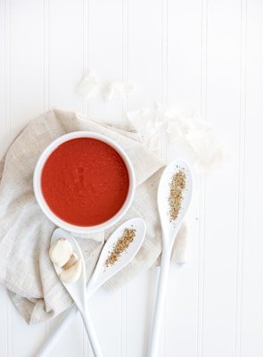 A flat lay composition featuring a white bowl filled with rich red tomato soup resting on a light-colored cloth. Next to the bowl, three white spoons are arranged artistically, each holding dried herbs. There are also three cloves of garlic placed on one of the spoons. The background is a clean, white wooden surface, enhancing the minimalist and fresh look of the setup.
