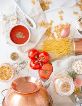 A selection of fresh and raw cooking ingredients are laid out on a white surface. There are tomatoes on the vine, a copper colander, garlic cloves, a shallot, and various types of pasta scattered around. Two white spoons contain herbs, and a small white bowl is filled with tomato sauce, sprinkled with additional herbs. A jar of spiral pasta and a glass container of spices are also present.