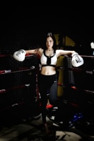 A woman stands confidently in a boxing ring, wearing white boxing gloves and athletic gear. The low lighting creates a dramatic atmosphere, highlighting her determined expression and athletic physique. The background is dark with hints of gym equipment visible.