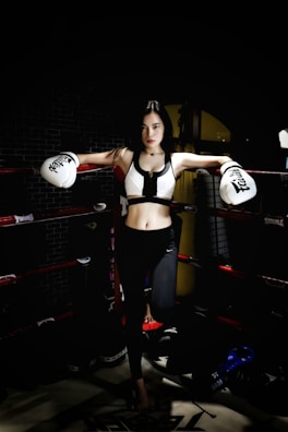 A woman stands confidently in a boxing ring, wearing white boxing gloves and athletic gear. The low lighting creates a dramatic atmosphere, highlighting her determined expression and athletic physique. The background is dark with hints of gym equipment visible.