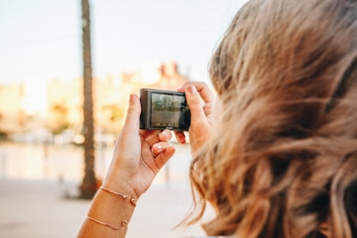 A person with light brown, wavy hair is seen from behind, holding a small digital camera, focusing on capturing an external scene in sunny weather. The background appears slightly blurred, with soft tones suggesting a leisurely outdoor environment.