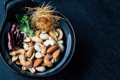 A black bowl contains a mixture of nuts and other ingredients. Cashews and almonds are prominently visible along with a few dark red dried chili peppers. There are also some green leaves and a nest of thin, straw-like strands in the bowl. The bowl rests on a dark surface.