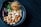 A black bowl contains a mixture of nuts and other ingredients. Cashews and almonds are prominently visible along with a few dark red dried chili peppers. There are also some green leaves and a nest of thin, straw-like strands in the bowl. The bowl rests on a dark surface.