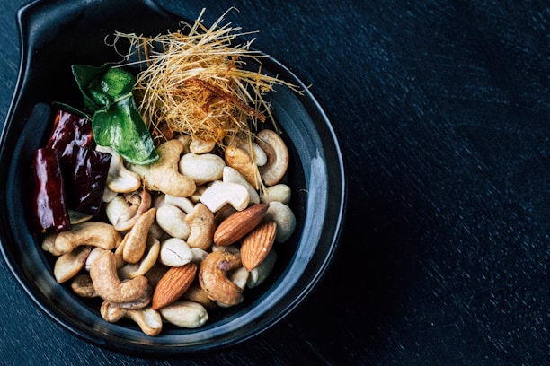 A black bowl contains a mixture of nuts and other ingredients. Cashews and almonds are prominently visible along with a few dark red dried chili peppers. There are also some green leaves and a nest of thin, straw-like strands in the bowl. The bowl rests on a dark surface.