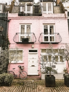 A charming pink brick townhouse with a quaint facade featuring white-framed windows and doors. Two narrow balconies with ornate metal railings adorn the upper floors. A bicycle with a wicker basket leans against the wall, surrounded by large potted plants. The cobblestone pavement adds to the cozy, rustic ambiance.