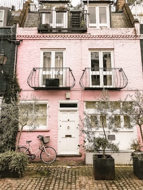A charming pink brick townhouse with a quaint facade featuring white-framed windows and doors. Two narrow balconies with ornate metal railings adorn the upper floors. A bicycle with a wicker basket leans against the wall, surrounded by large potted plants. The cobblestone pavement adds to the cozy, rustic ambiance.