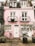 A charming pink brick townhouse with a quaint facade featuring white-framed windows and doors. Two narrow balconies with ornate metal railings adorn the upper floors. A bicycle with a wicker basket leans against the wall, surrounded by large potted plants. The cobblestone pavement adds to the cozy, rustic ambiance.