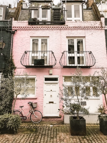 A charming pink brick townhouse with a quaint facade featuring white-framed windows and doors. Two narrow balconies with ornate metal railings adorn the upper floors. A bicycle with a wicker basket leans against the wall, surrounded by large potted plants. The cobblestone pavement adds to the cozy, rustic ambiance.