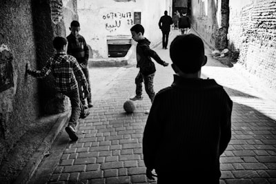 Four children are playing soccer on a cobblestone street, surrounded by old walls with graffiti. The scene is captured in black and white, with a focus on their movement and interaction with the ball.
