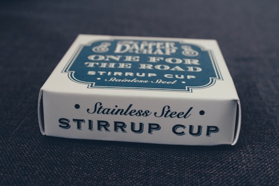 A white rectangular box sits on a dark surface. The front of the box is labeled with the text 'Stainless Steel STIRRUP CUP' in a bold, dark font. The top of the box also contains text, but it is partially obscured, with 'One for the Road' and 'Stainless Steel' visible.