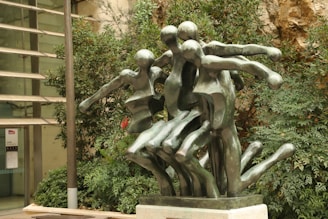 Several abstract human-like figures crafted from bronze are intertwined in a dynamic artistic sculpture. The setting includes lush greenery and a modern architectural background.