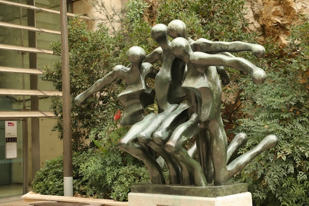 Several abstract human-like figures crafted from bronze are intertwined in a dynamic artistic sculpture. The setting includes lush greenery and a modern architectural background.