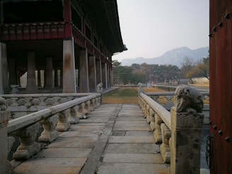 A traditional architectural structure features a stone pathway with ornate railings leading to a large building supported by multiple columns. The surrounding area consists of neatly trimmed grass and trees, with mountains visible in the background under a pale sky.