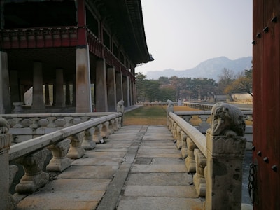 A traditional architectural structure features a stone pathway with ornate railings leading to a large building supported by multiple columns. The surrounding area consists of neatly trimmed grass and trees, with mountains visible in the background under a pale sky.