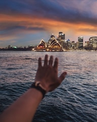 A hand is extended towards the Sydney Opera House across a body of water, with a city skyline in the background under a dramatic, colorful sky at dusk.