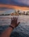 A hand is extended towards the Sydney Opera House across a body of water, with a city skyline in the background under a dramatic, colorful sky at dusk.
