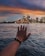 A hand is extended towards the Sydney Opera House across a body of water, with a city skyline in the background under a dramatic, colorful sky at dusk.