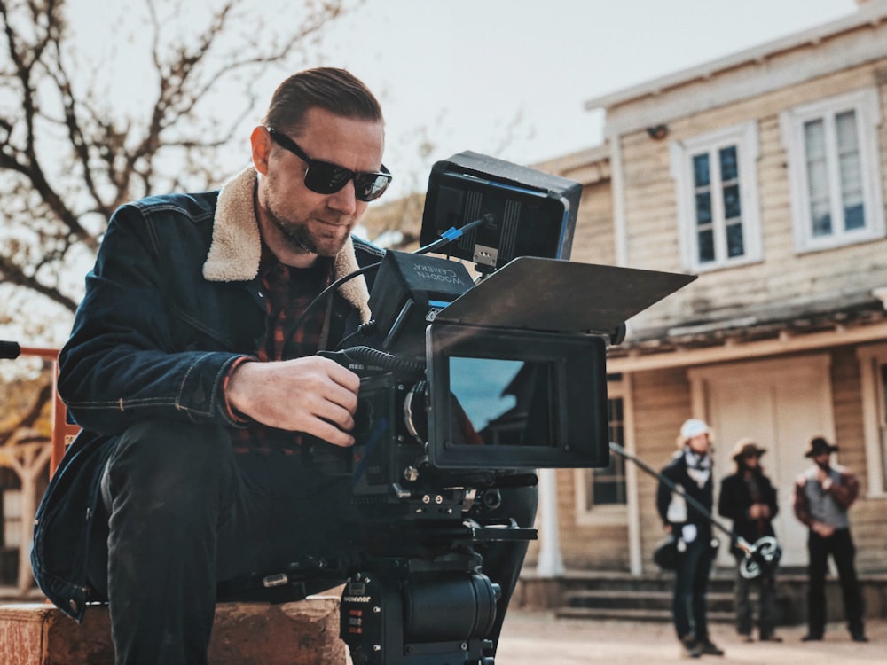 man sitting on bench near house while holding camera during daytime