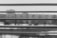 Horizontal blinds in the foreground partially obscure the view of an out-of-focus cityscape in black and white. The scene beyond appears to be an urban environment with trees and water, creating a soft, blurred contrast to the sharp lines of the blinds.