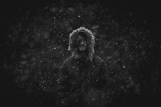 A person wearing a hooded parka stands in the middle of a snowy scene with snowflakes falling around them. The image is in black and white, creating a moody and atmospheric effect. The hood is lined with fur, framing the person's face.