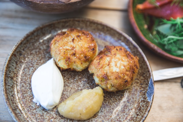 Two golden-brown potato pancakes are served on a rustic ceramic plate alongside a dollop of sour cream and a serving of apple sauce.