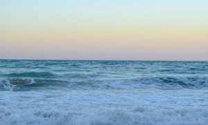 A serene ocean scene with gentle waves approaching the shore under a clear sky. The horizon is visible, dividing the sea and sky with a soft gradient of colors. The water is a vibrant mix of blues and greens, and the sky has a faint orange hue suggesting a sunrise or sunset.