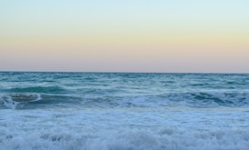 A serene ocean scene with gentle waves approaching the shore under a clear sky. The horizon is visible, dividing the sea and sky with a soft gradient of colors. The water is a vibrant mix of blues and greens, and the sky has a faint orange hue suggesting a sunrise or sunset.