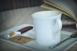 A white ceramic coffee mug sits on a tray beside a spoon, a sugar packet, a cigarette, and a book with open pages.