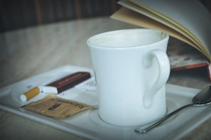 A white ceramic coffee mug sits on a tray beside a spoon, a sugar packet, a cigarette, and a book with open pages.