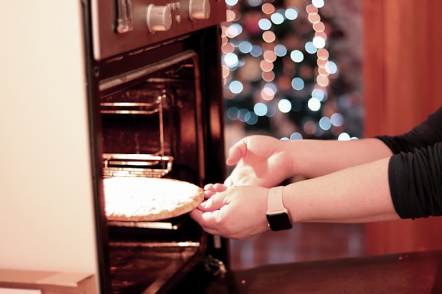 A person placing or removing a pizza from a conventional oven. The image features a blurred background with multicolored bokeh lights, suggesting a festive or cozy setting. The focus is on the person's hands and the pizza, with an emphasis on the open oven door and control knobs.