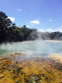 A landscape featuring a geothermal hot spring with steam rising from the surface. The foreground shows a mineral-rich area with earthy tones. Lush green trees are in the background under a clear blue sky.