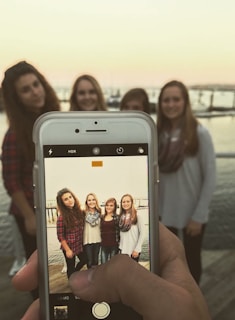 A hand is holding a smartphone, capturing a photo of four women standing together on a wooden dock with a body of water and boats in the background. The screen shows a clearer image of the women than the blurred background, highlighting their casual attire and scarves.
