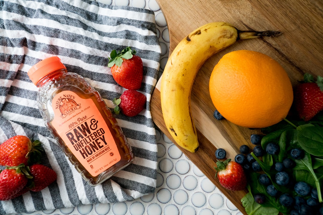 raw and unfiltered honey bottle between strawberries with blueberries, citrus, and banana fruits nearby