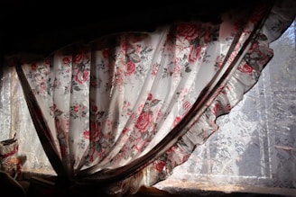 A floral curtain with intricate patterns of red and pink roses hangs partially open, allowing sunlight to filter through. The lace underneath adds a delicate touch, complementing the romantic and vintage aesthetic of the fabric.
