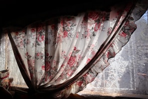 A floral curtain with intricate patterns of red and pink roses hangs partially open, allowing sunlight to filter through. The lace underneath adds a delicate touch, complementing the romantic and vintage aesthetic of the fabric.
