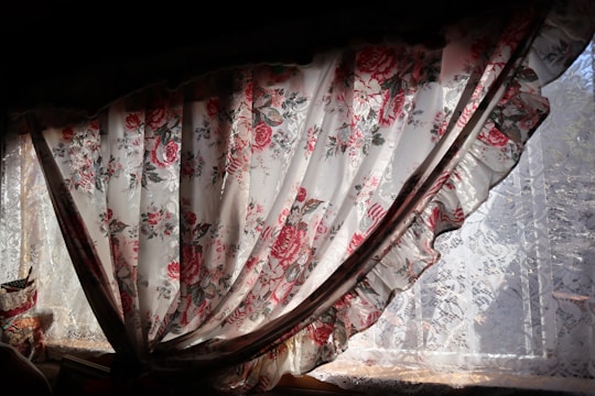 A floral curtain with intricate patterns of red and pink roses hangs partially open, allowing sunlight to filter through. The lace underneath adds a delicate touch, complementing the romantic and vintage aesthetic of the fabric.