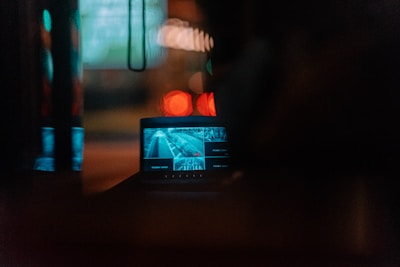 A dark room with a monitor displaying a blurred image. The screen contains a scene of a road or street, possibly showing a surveillance or security footage. Soft lights cast around the scene create a moody atmosphere.