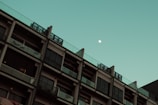 A multi-story building with modern architecture is seen against a clear sky with a visible moon. The building has numerous windows and balconies, and each level showcases a uniform design. The moon is positioned in the upper right portion of the sky, adding an element of tranquility to the scene.