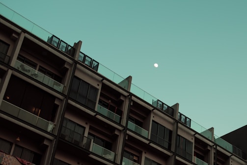 A multi-story building with modern architecture is seen against a clear sky with a visible moon. The building has numerous windows and balconies, and each level showcases a uniform design. The moon is positioned in the upper right portion of the sky, adding an element of tranquility to the scene.