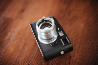 A vintage-style camera with a black body and a prominent silver lens placed on a wooden surface. The image is artfully blurred, creating a soft focus on the camera.